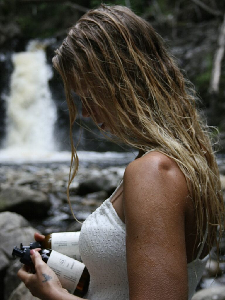 kvinne holder rå skincare sjampo og balsam i hånden ved en foss. håret er vått, naturlig og rent