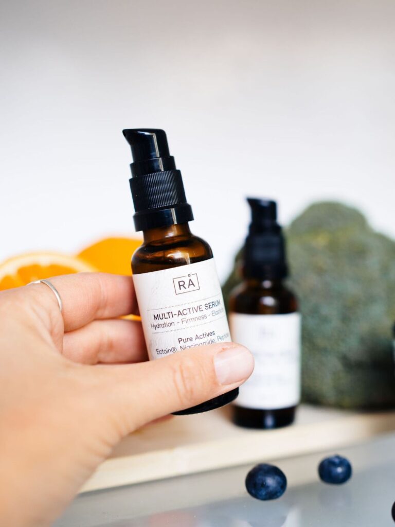 Multi-Active Serum fra RÅ Skincare holdes i hånden foran friske blåbær, frukt og grønne ingredienser.