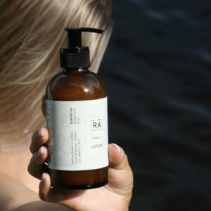 Grøde lotion holdes opp. I bakgrunnen ser du en dame med langt hår som blåser i vinden og havet i bakgrunnen