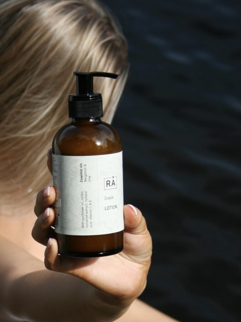 Grøde lotion holdes opp. I bakgrunnen ser du en dame med langt hår som blåser i vinden og havet i bakgrunnen