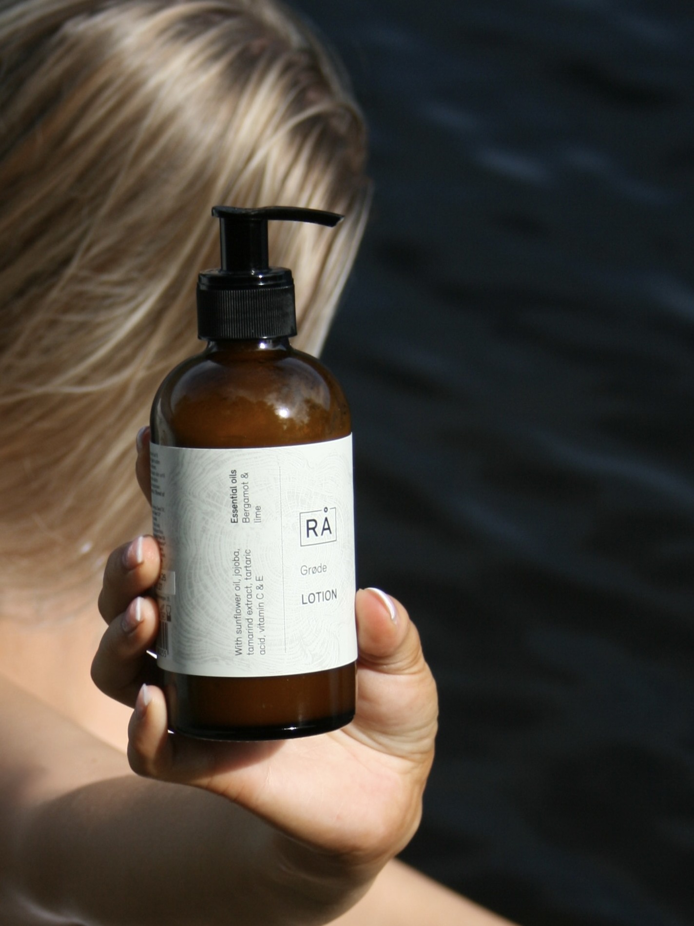 Grøde lotion holdes opp. I bakgrunnen ser du en dame med langt hår som blåser i vinden og havet i bakgrunnen