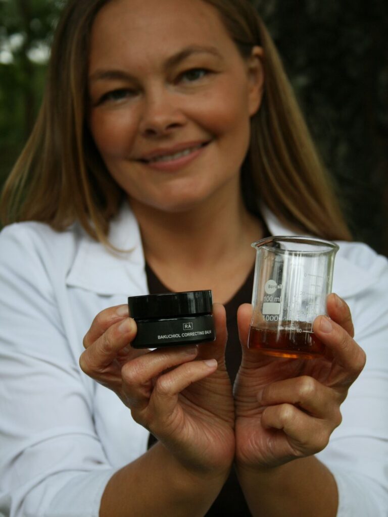 Amanda holder frem Bakuchiol ingrediensen i et labglass sammen med rå bakuchiol correcting balm for å vise produktet og ingrediensene. Hun står ute i labfrakk
