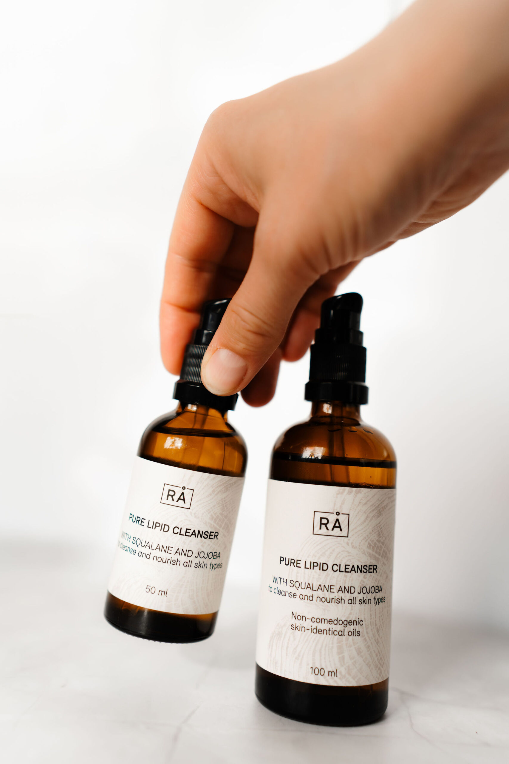 Pure lipid cleanser 50 ml og 100 ml vises sammen. En hånd holder reisestørrelsen på 50 ml.