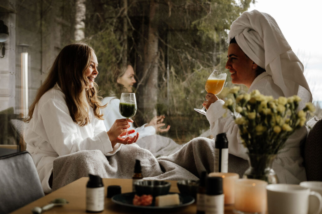 To damer drikker smudi i tretopphytte med utsikt ut over skogen De sitter ved et bord med hudpleie og tapas