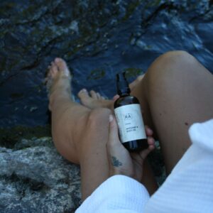 Kvinne i hvit morgenkåpe sitter på en stein ved vannkanten og holder RÅ Grøde massage and body oil i hendene.