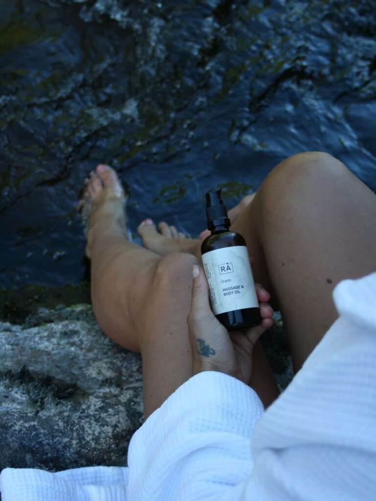 Woman in white bathrobe sitting on a rock by the water's edge holding RÅ Grøde massage and body oil in her hands.