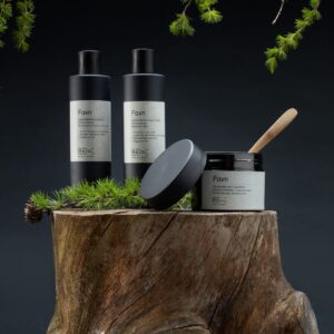 RÅ Favn hårpleieserie med sjampo, balsam og hårkur plassert på en trestubbe omgitt av grankvister.