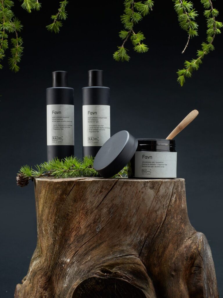 RÅ Favn hårpleieserie med sjampo, balsam og hårkur plassert på en trestubbe omgitt av grankvister.